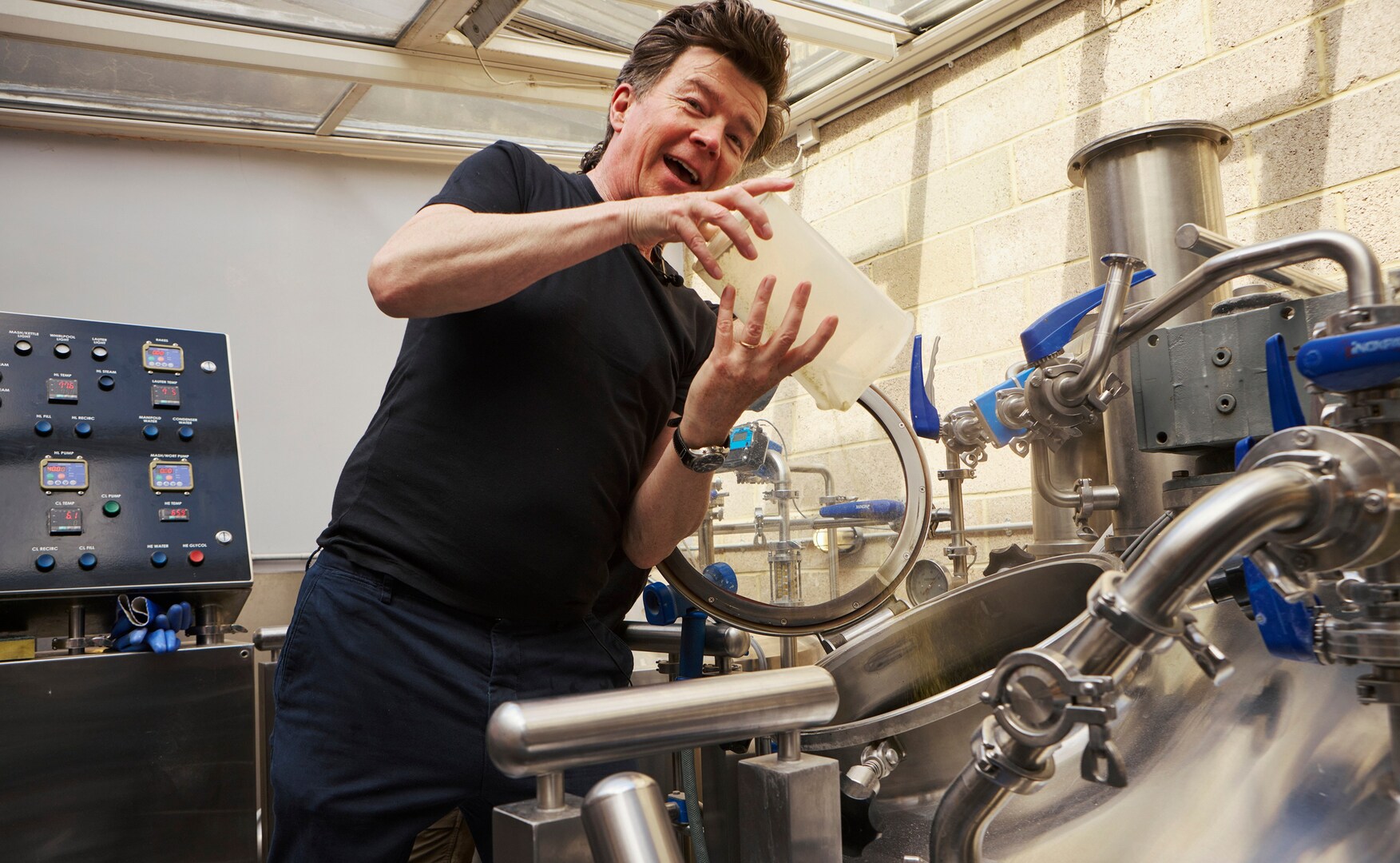 Rick Astley adding ingredients to a large metal brewing tank.